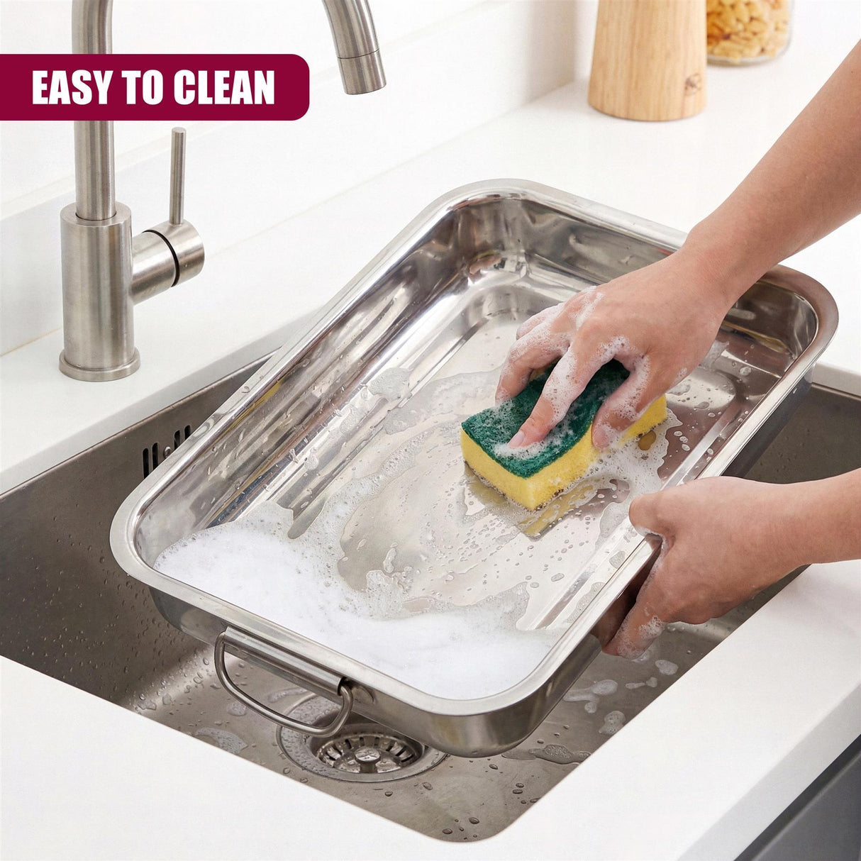 A hand washes a rectangular stainless steel tray containing suds in a kitchen sink.