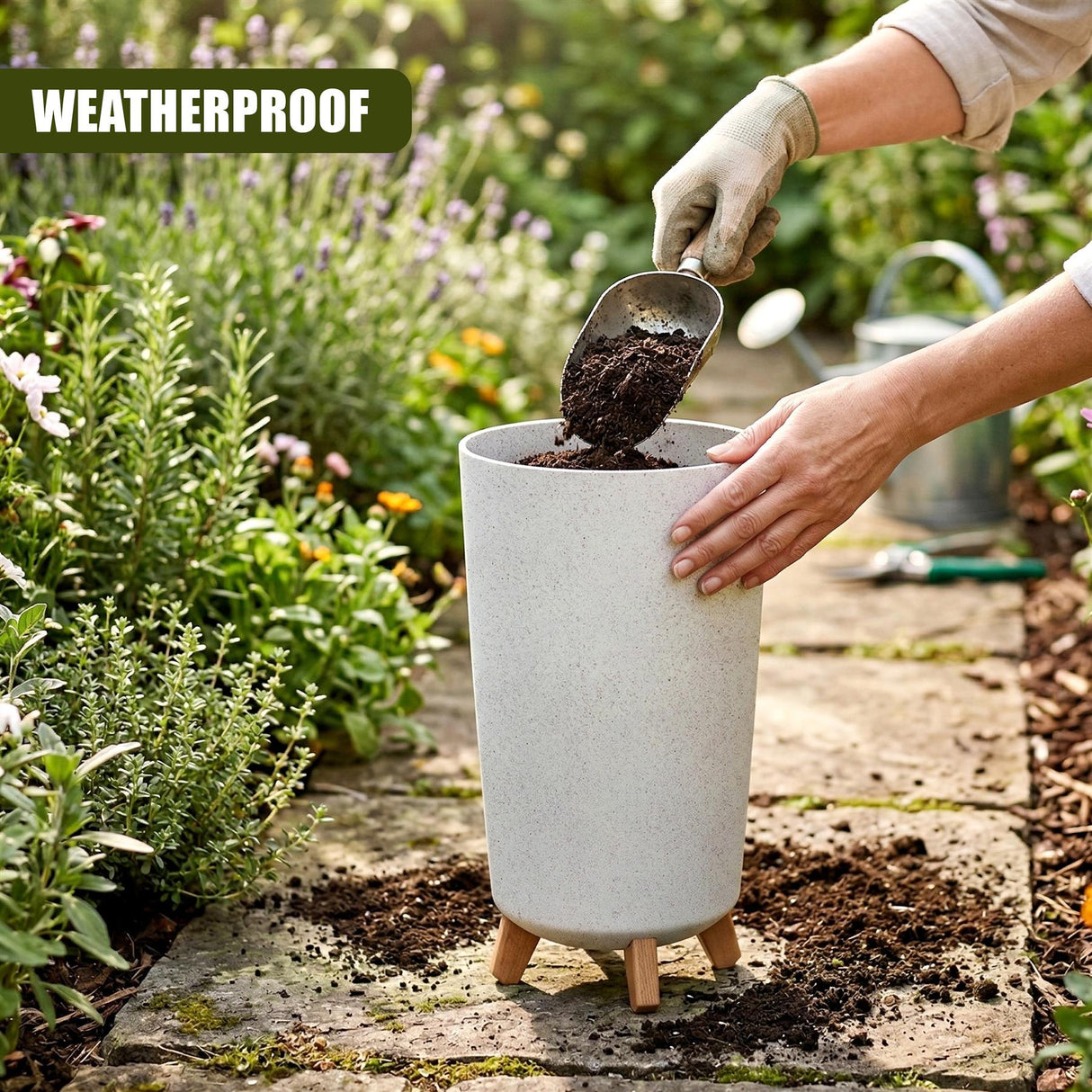 A gray, legged flower pot receives dark soil from a hand using a small garden scoop.