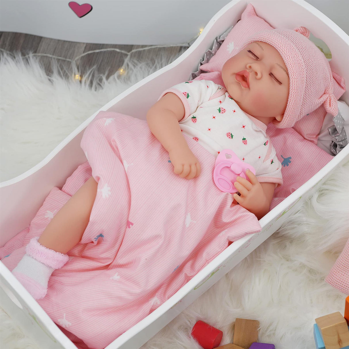 A doll rests in a pink wooden crib with a striped blanket and matching pink hat.