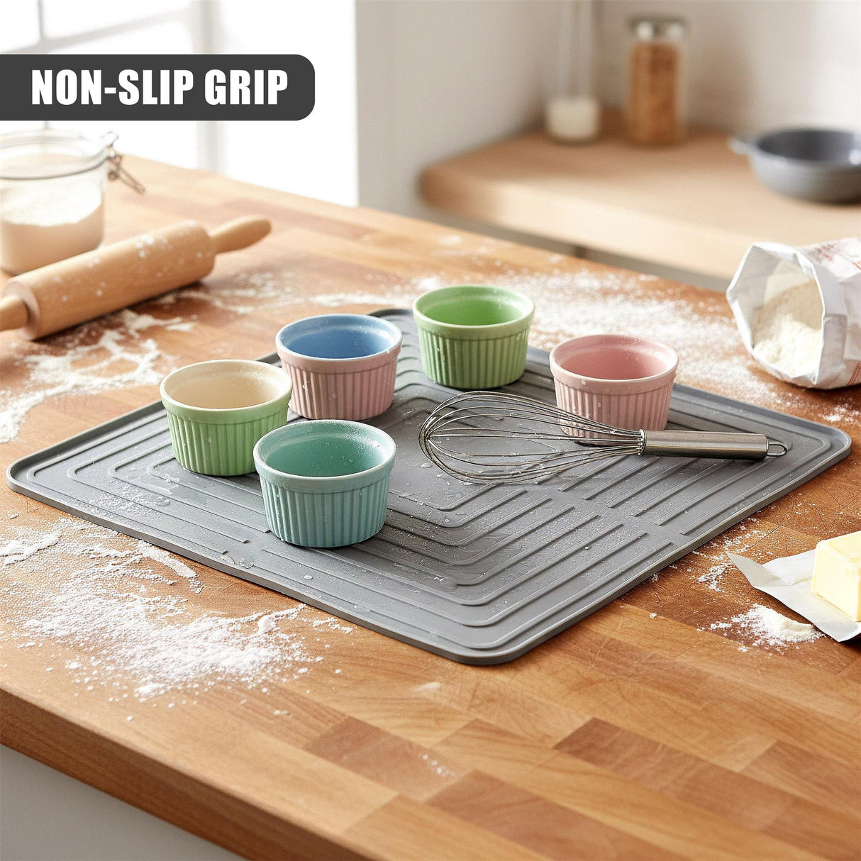 A grey drying mat holds multiple colorful cups and a whisk, surrounded by baking flour on wood.