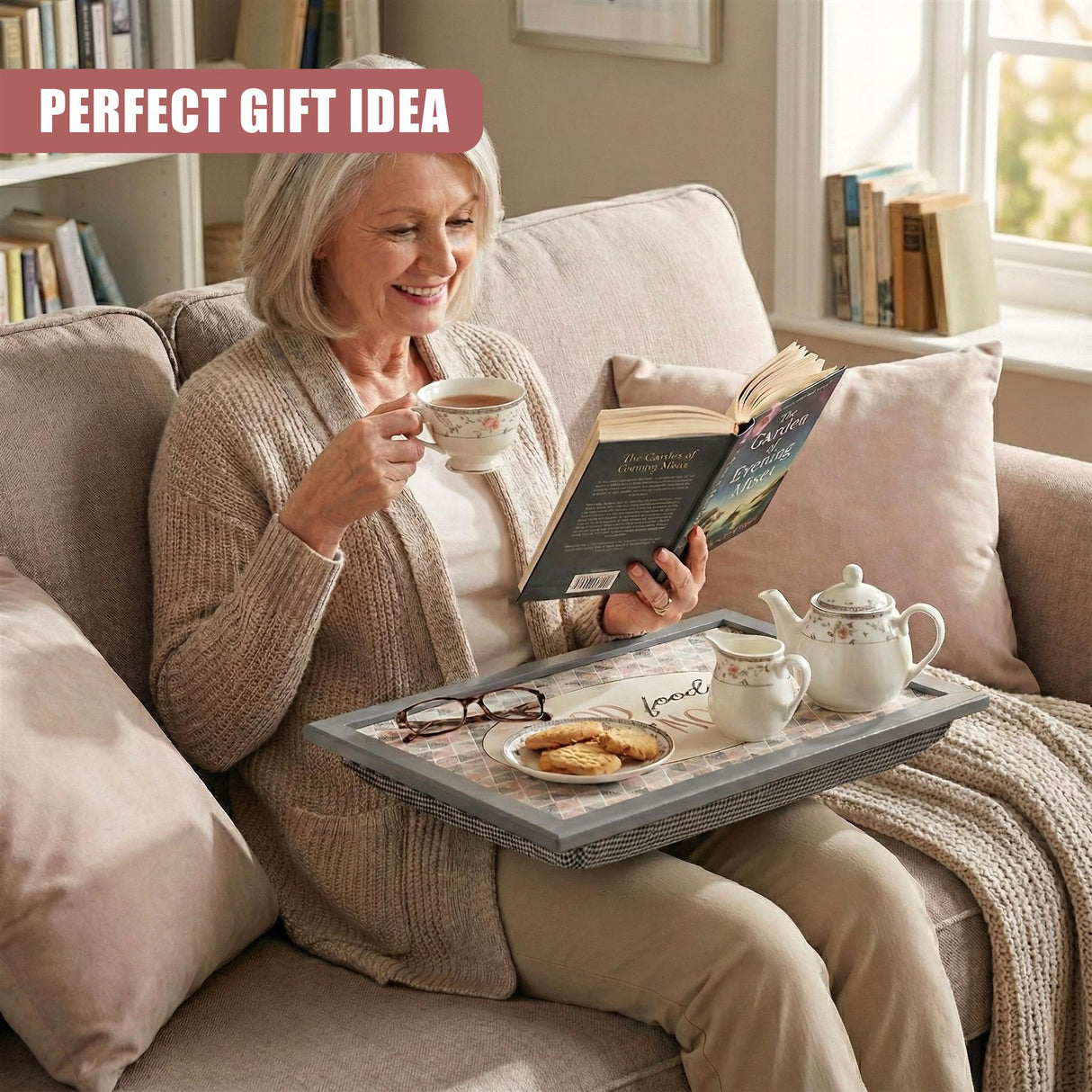 A lap tray holding snacks, a book, and a cream pitcher used by a woman sitting on a couch.