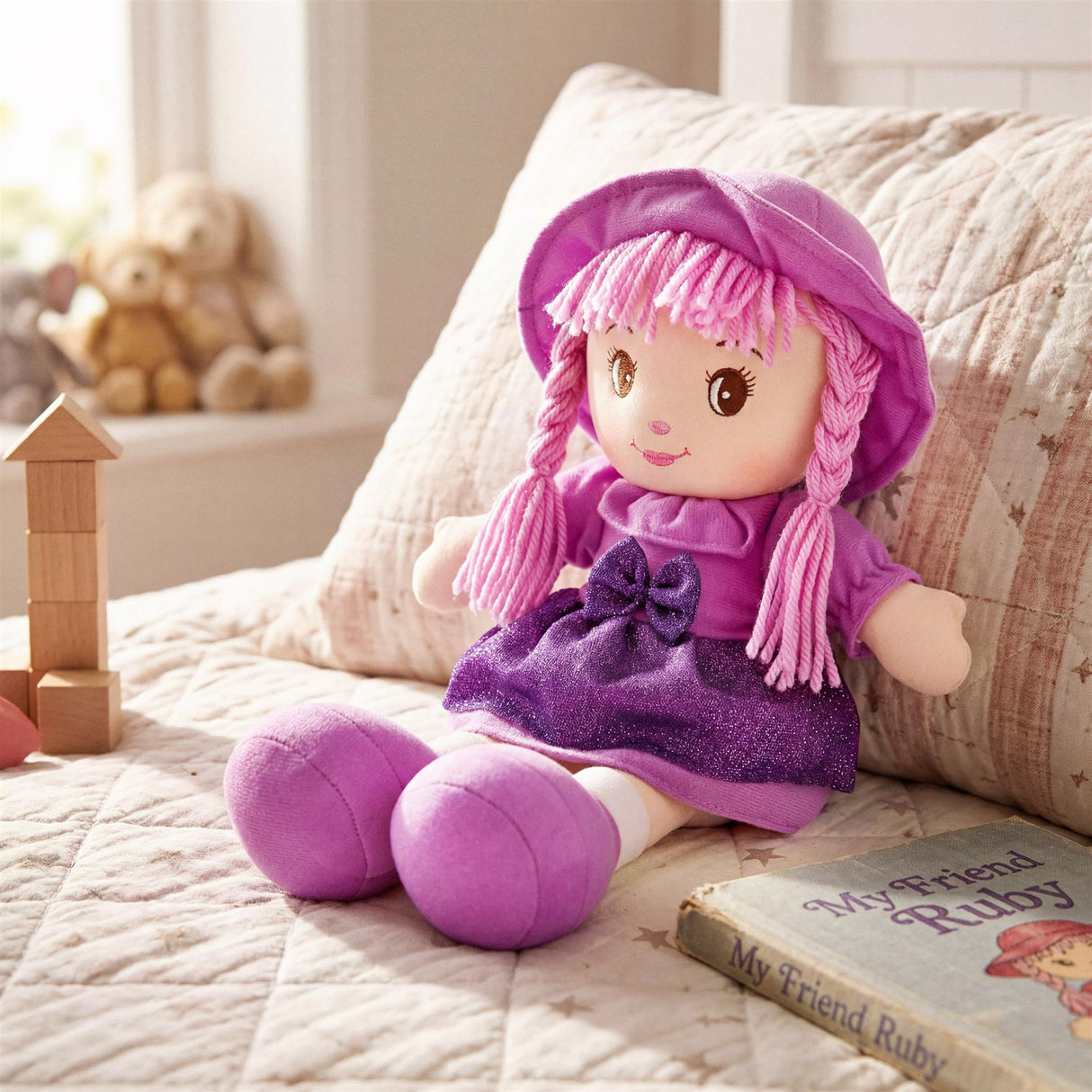 Rag doll Ruby, wearing a purple hat and dress, rests on a bed near stacked wooden blocks and a book.