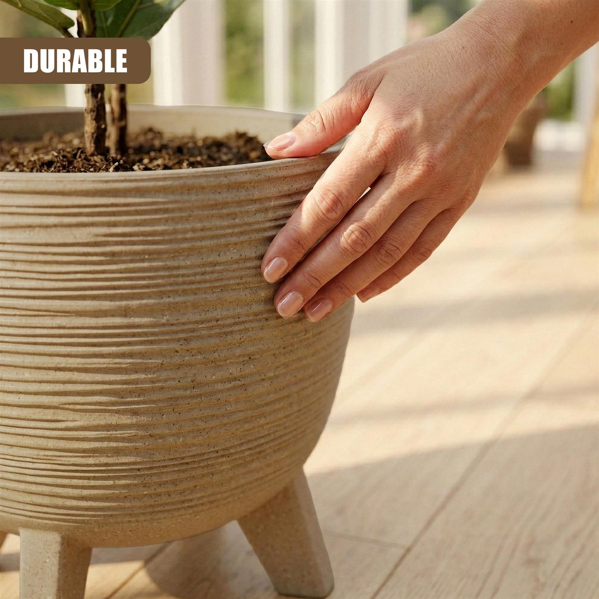 Large, ribbed brown flower pot on legs contains soil and a small plant. A hand rests near the rim.