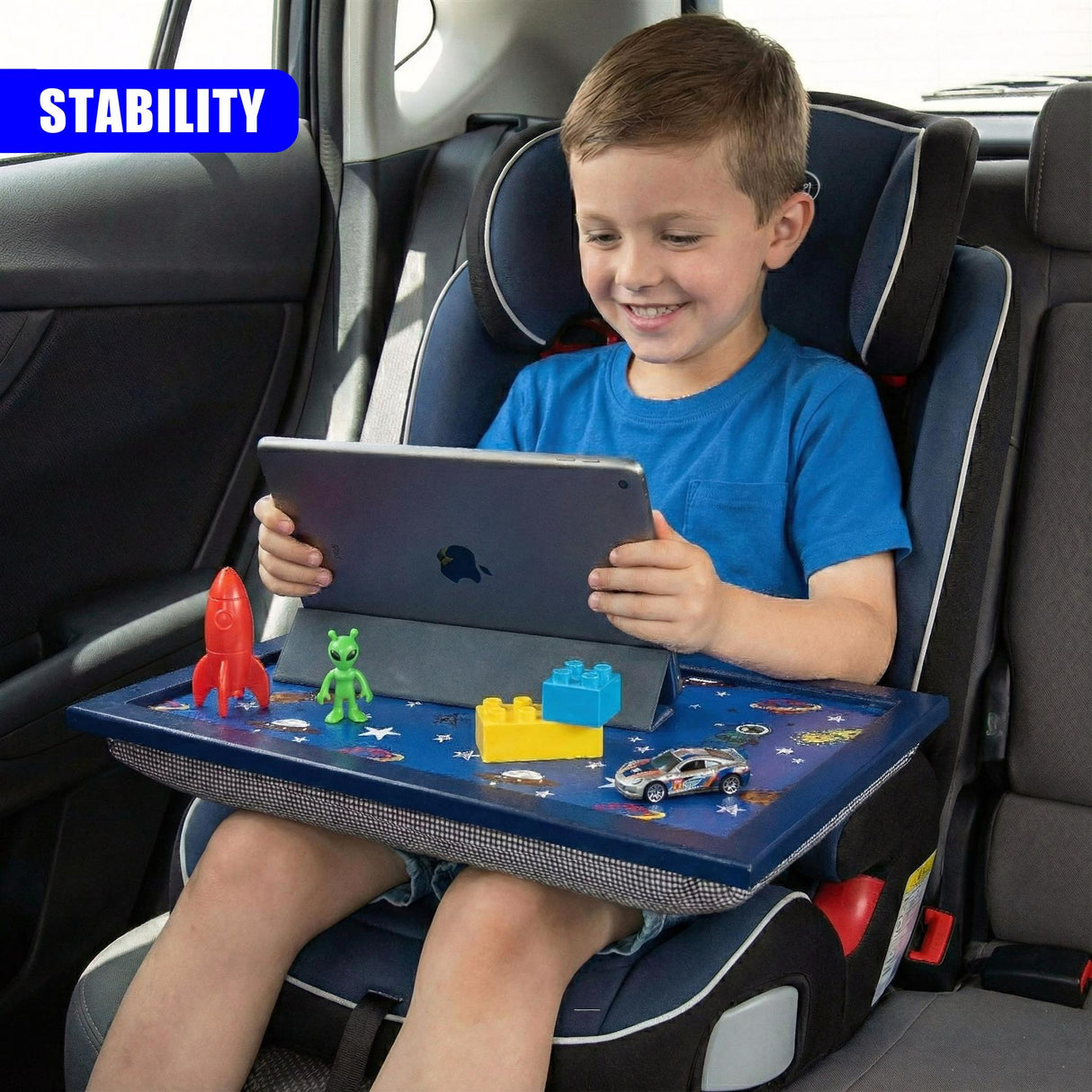 A blue lap tray attached to a car seat holds a tablet and various plastic toys for a young boy.