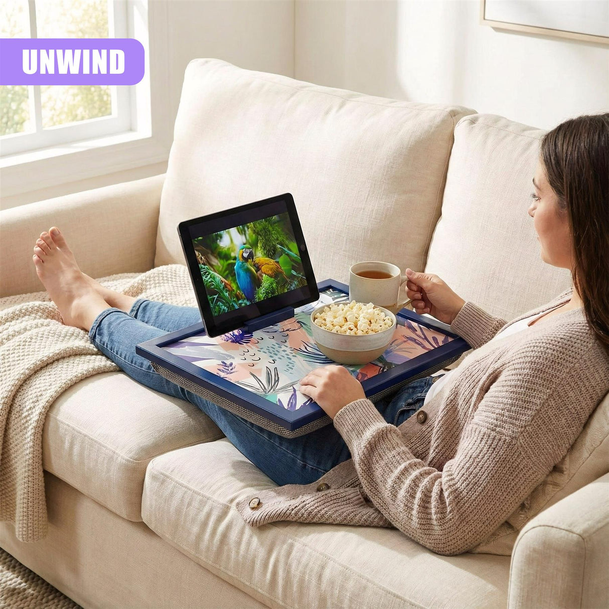 A woman is sitting on a couch, holding a tray with a tablet, a bowl of popcorn, and a cup of tea. The tray has a colorful design and a built-in stand for the tablet. The woman is watching a movie on the tablet, and there is a window with a view of a tree in the background. The room is bright and sunny, with a white wall and a few plants. The woman is wearing a cozy sweater and jeans, and her feet are covered with a blanket. - UKBuyZone