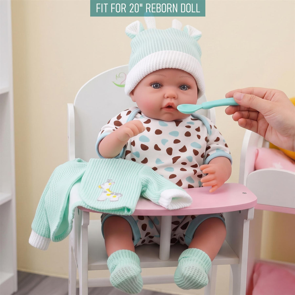 A reborn doll in mint and spotty clothes is sitting at a table while being fed by a hand.