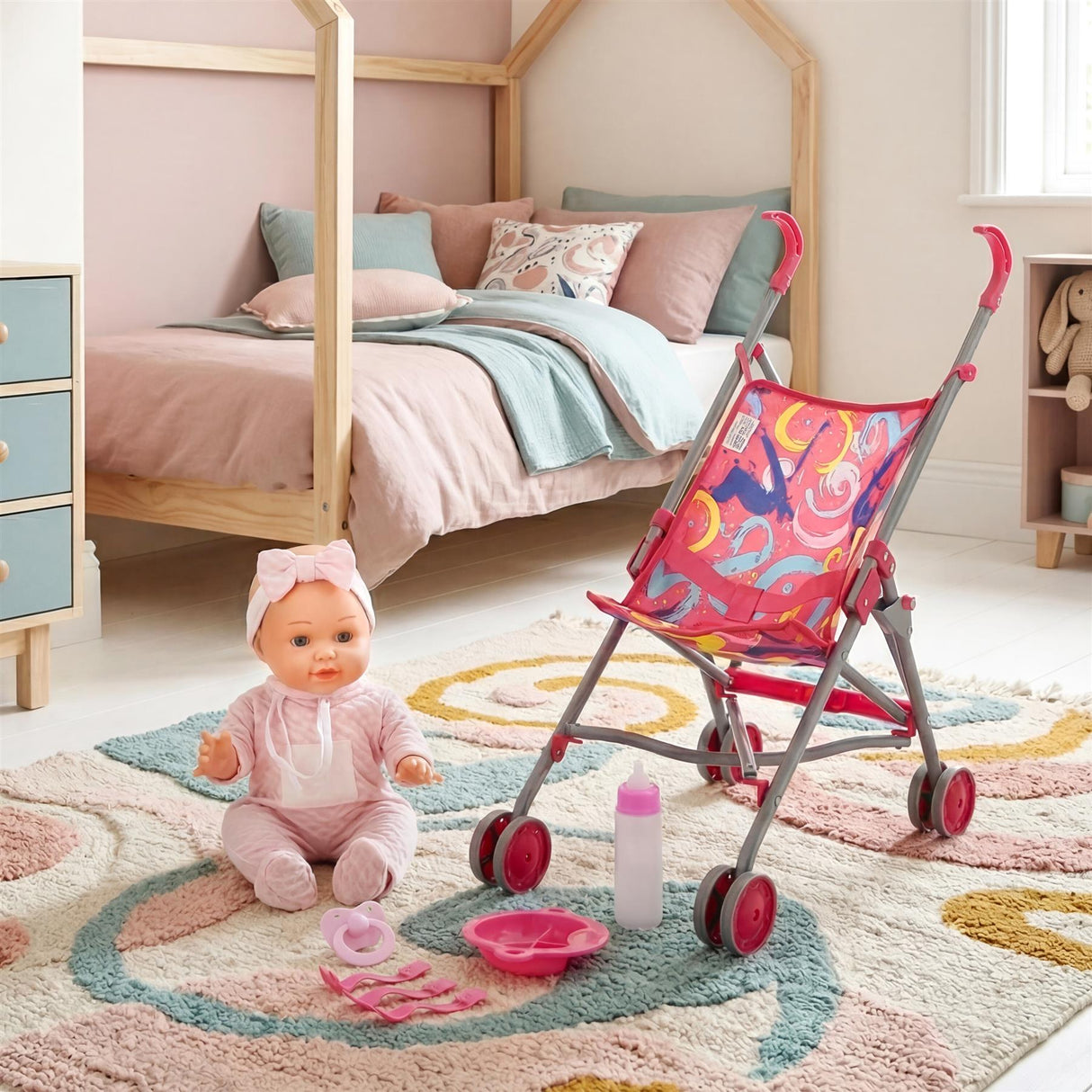 Doll with pink outfit sits beside a patterned red stroller and feeding accessories set.