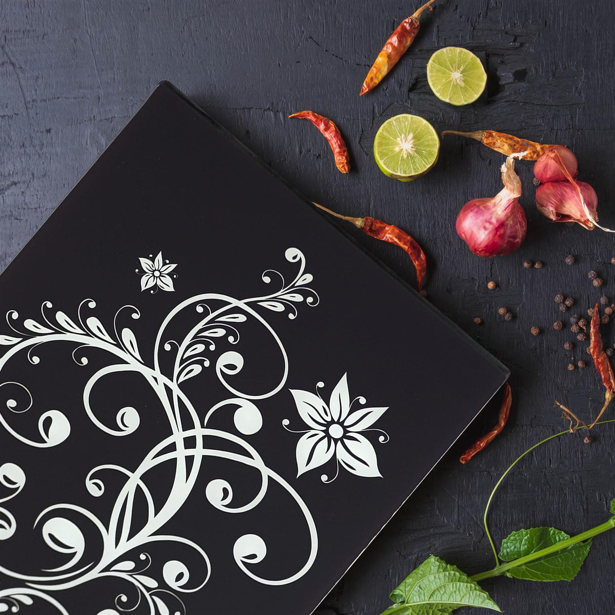 A black, rectangular cutting board with a white, floral design, surrounded by various ingredients like a lime, chili peppers, and onions. - UKBuyZone