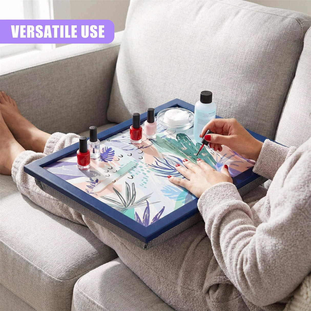 A woman sits on a couch, painting her nails on a tray with various nail polish colors and a nail polish remover. The tray is a dark blue, rectangular box with a removable tray top. The top has a colorful, abstract design with a mix of plants and abstract shapes. The woman is wearing a light gray robe and has her feet up on the couch. She is painting her nails on the tray, which is placed on her lap. - UKBuyZone