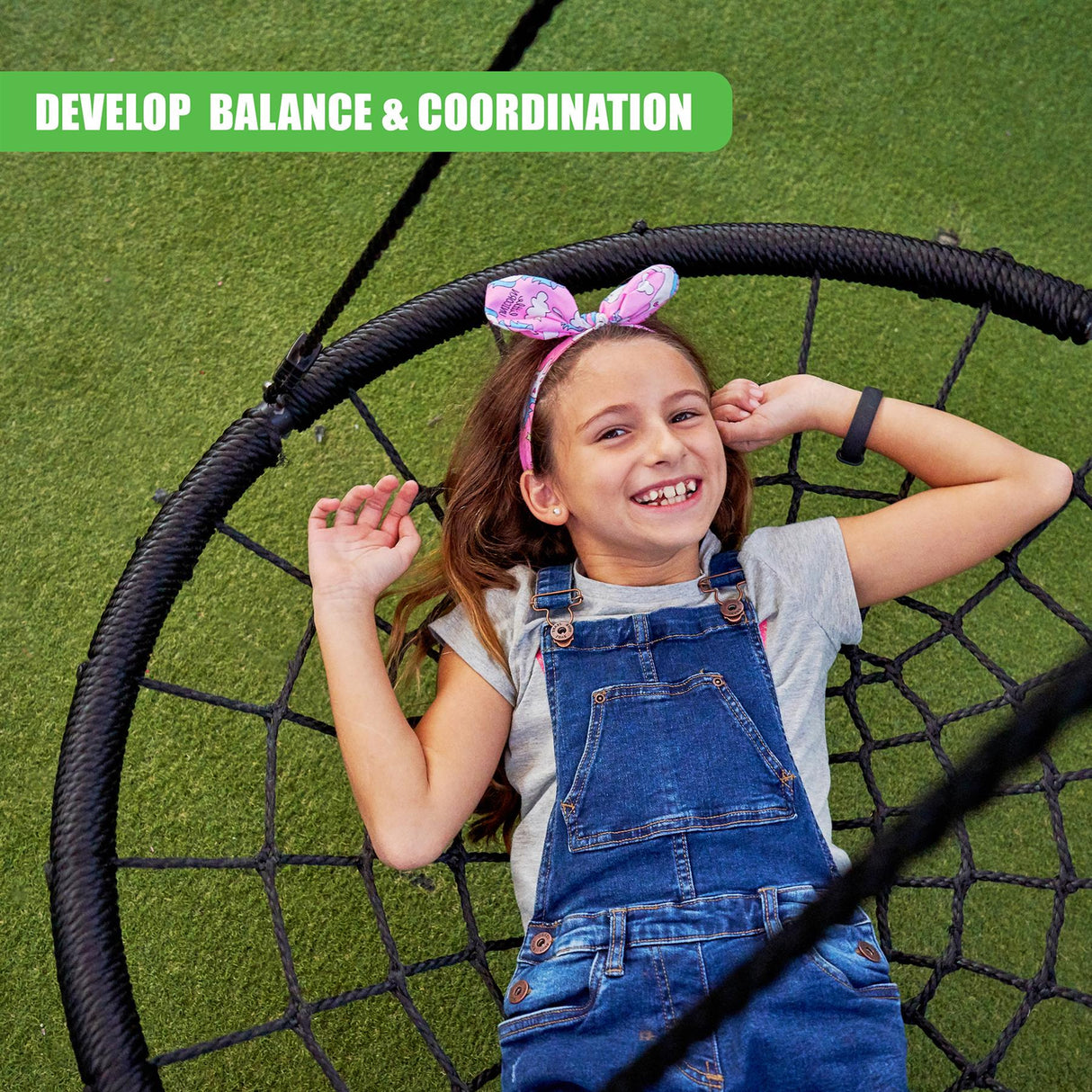 A smiling young girl in blue overalls sits on a dark, woven backyard swing set.