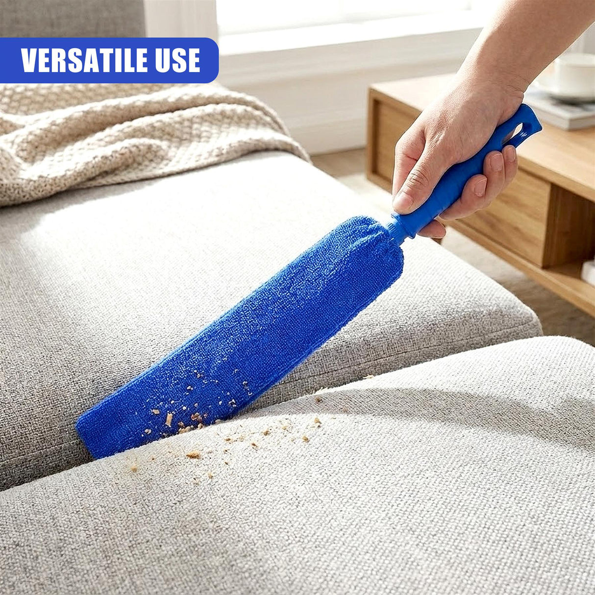 Hands use a blue duster to clean dust from a light gray upholstered sofa.