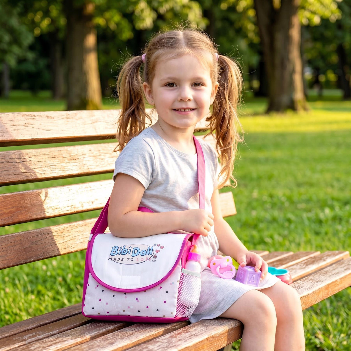 A young girl sits on a wooden bench with a pink patterned doll changing bag and accessories.