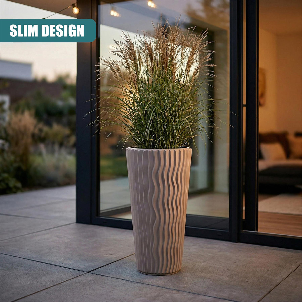 Tall, ribbed, light brown flower pot containing voluminous, feathery grasses placed near glass doors.