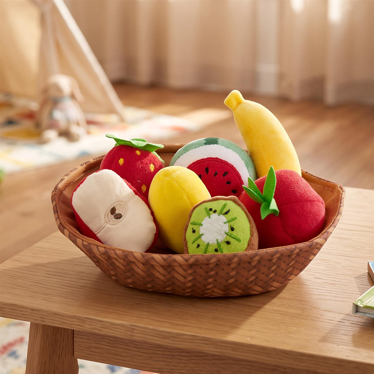 A brown woven fabric basket filled with seven soft plush fruits including a banana, strawberry, watermelon, kiwi, apple and