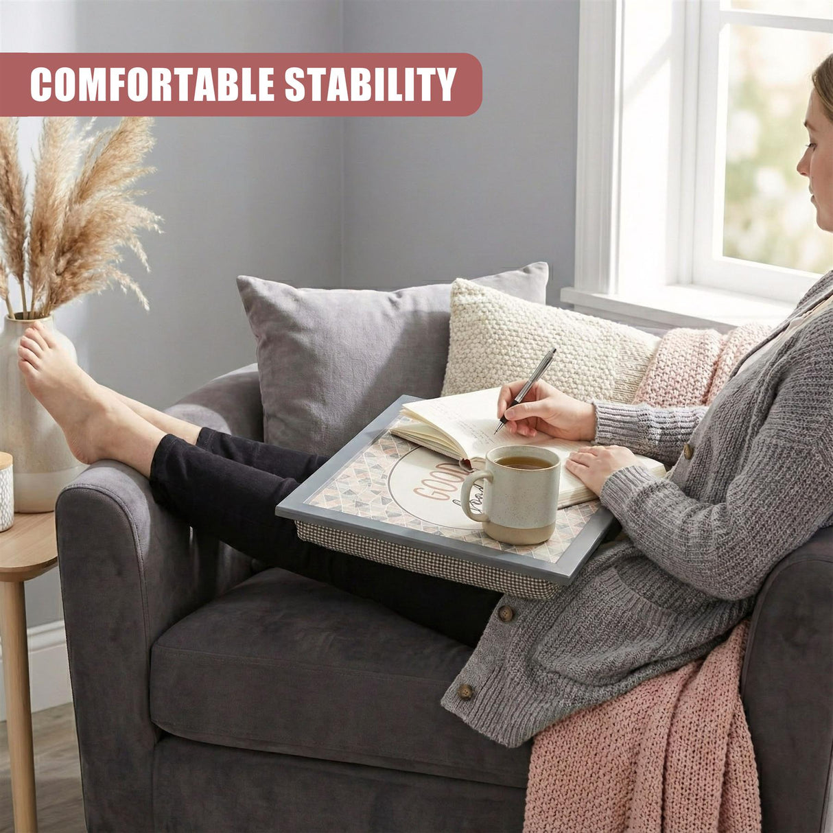 Rectangular patterned lap tray holding a mug and open book on a gray sofa next to a person.