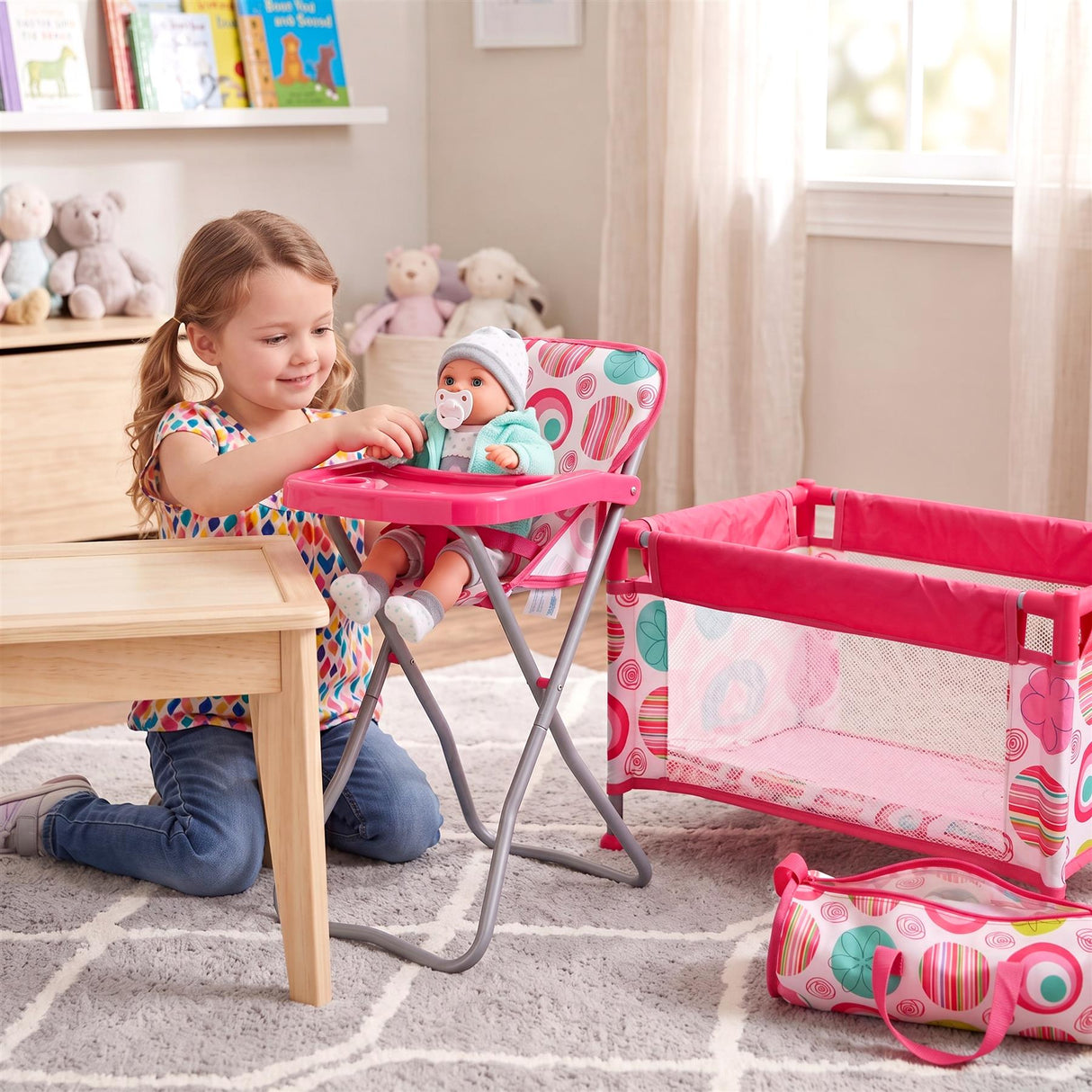 Pink baby cot, high chair, small wooden table, and patterned travel bag displayed in a staged child's room.
