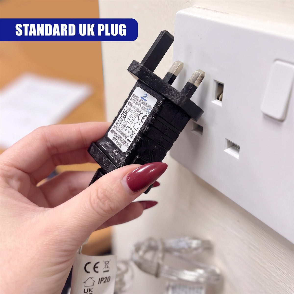 A hand holds a black battery eliminator adapter with a standard UK plug inserted into a white wall socket