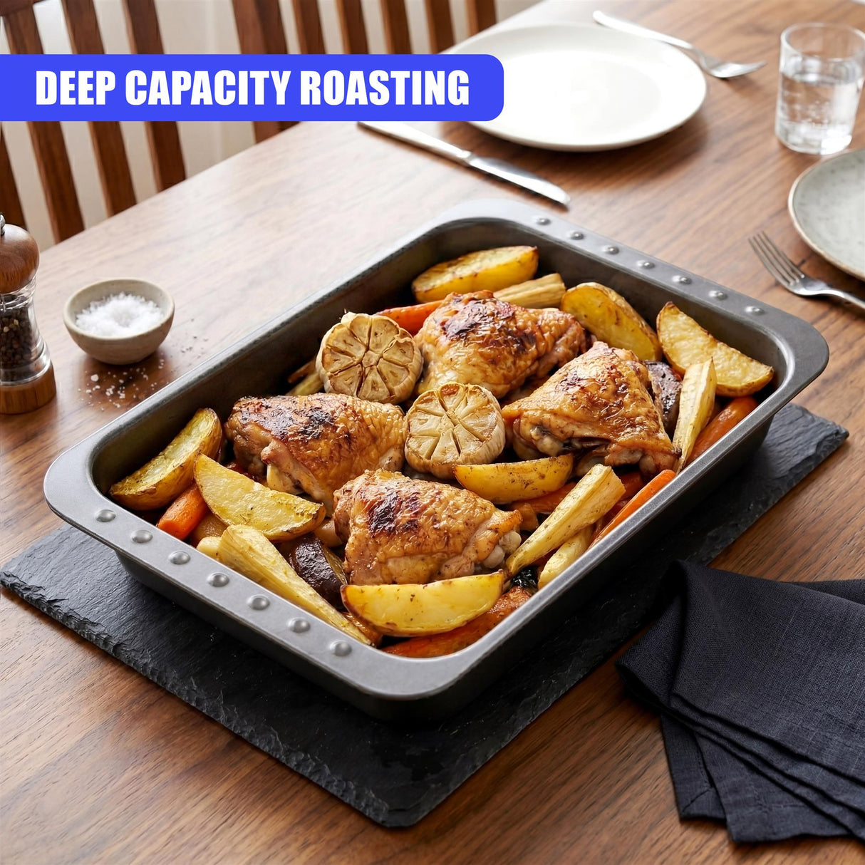 A dark metal roasting tin holds roasted chicken and sliced root vegetables on a black serving board.