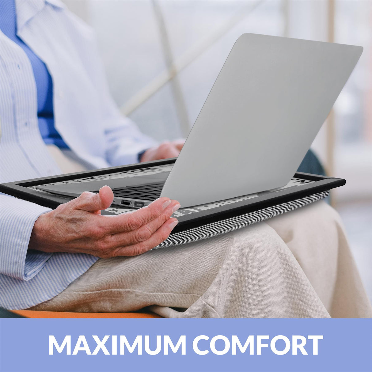 A person sits on a couch, holding a laptop on their lap, with a laptop stand underneath. The laptop is on a tray, and the person's hands are on the keyboard. The background is blurred, with a window visible in the distance. The person is wearing a blue shirt and a white jacket, and their hands are on the keyboard. The laptop stand is black and has a mesh design, and the laptop is a silver MacBook. - UKBuyZone