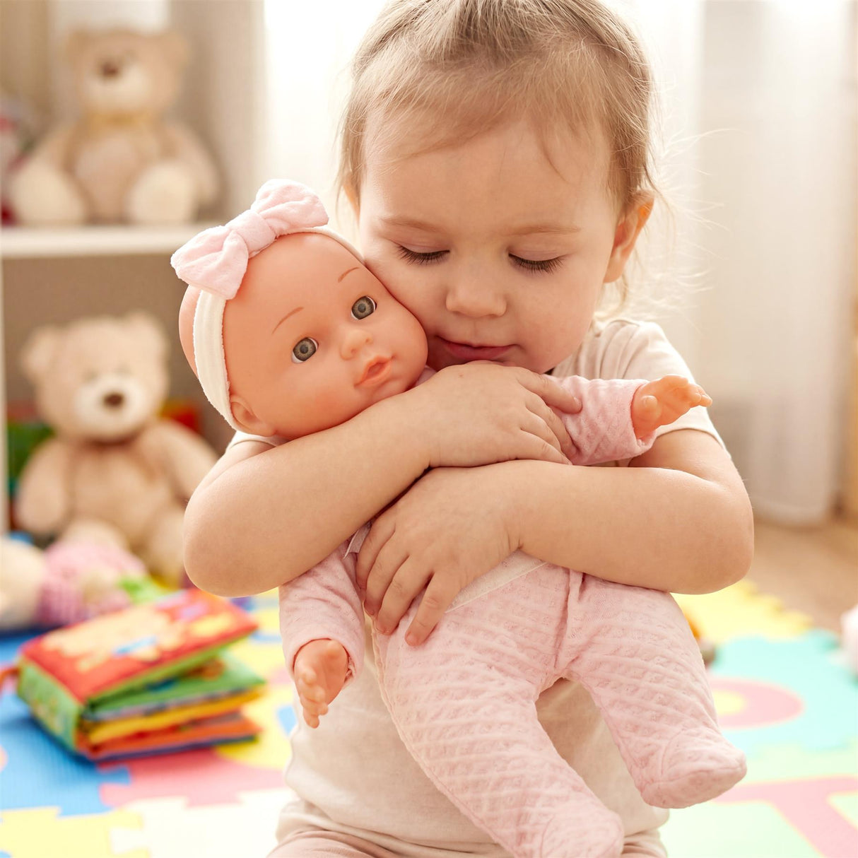 A toddler hugs a baby doll wearing a pink outfit, with plush toys and books visible in the background.