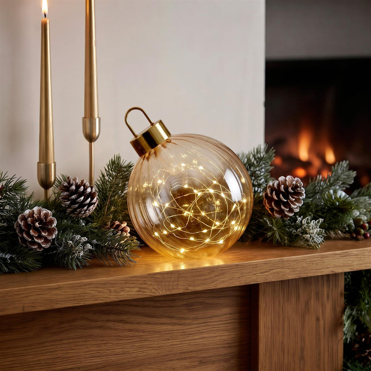 A large amber glass bauble with a gold top and internal fairy lights sits on a wooden mantelpiece decorated with pine
