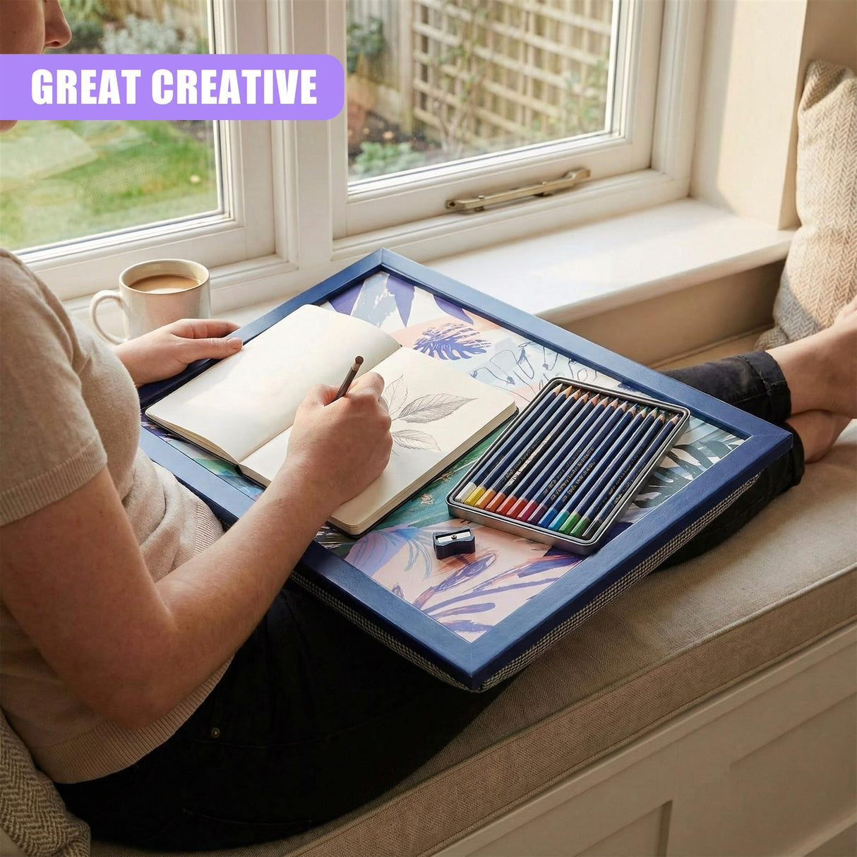 A woman sits on a window seat, surrounded by a tray of art supplies. She holds a sketchbook and pencil, while her feet are on a cushion. The tray is filled with a book, pencils, and a cup of coffee. A window is visible in the background, with a garden outside. The woman's face is not visible, but her hands and feet are. The scene is set in a peaceful, natural setting. - UKBuyZone