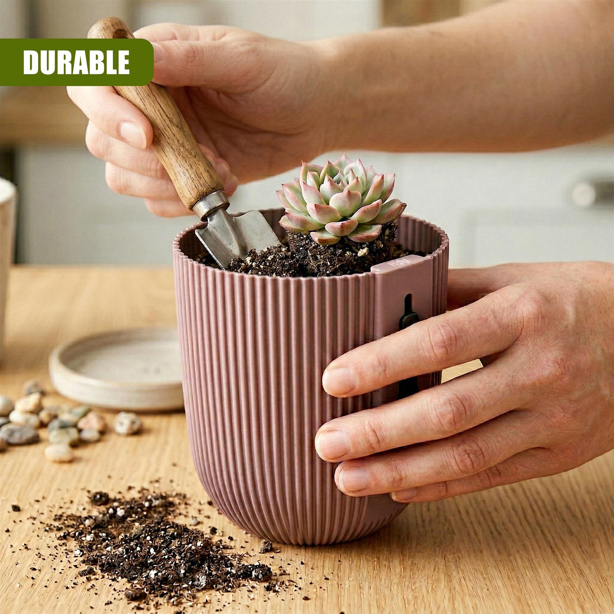 Hands planting a small succulent into a ribbed pink planter using a gardening trowel on a wooden surface.