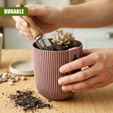 Hands planting a small succulent into a ribbed pink planter using a gardening trowel on a wooden surface.