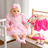 Large doll wears pink clothing near folded patterned clothes, shorts, and assorted baby shoes.