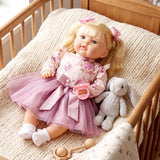 A blonde doll in a pink dress lies in a crib next to a stuffed bunny and wooden rattle.