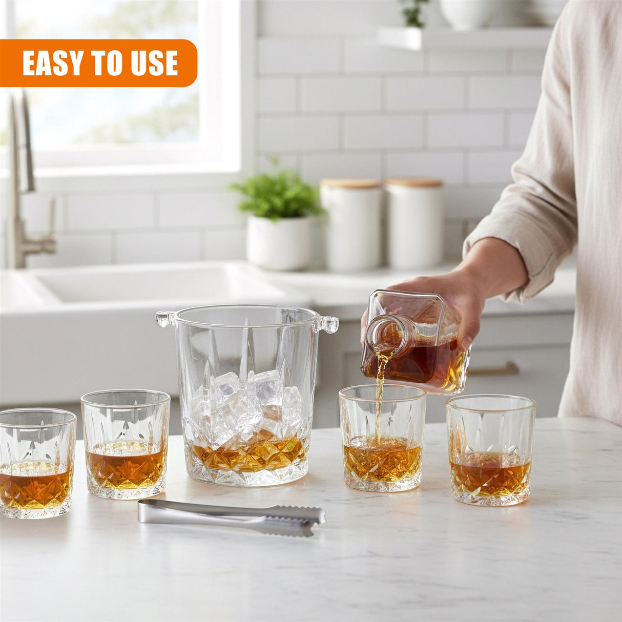A hand pours amber liquor from a glass pitcher into whiskey glasses next to an ice bucket.