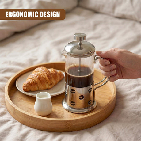 Hand holding a metal coffee pot over a wooden tray with croissant and creamer.