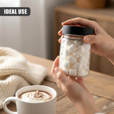 A hand holds a clear glass jar of white cubes next to a white mug containing a beverage.