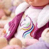 Doll wears a purple jacket with a white collar and a cream shirt displaying an embroidered unicorn.