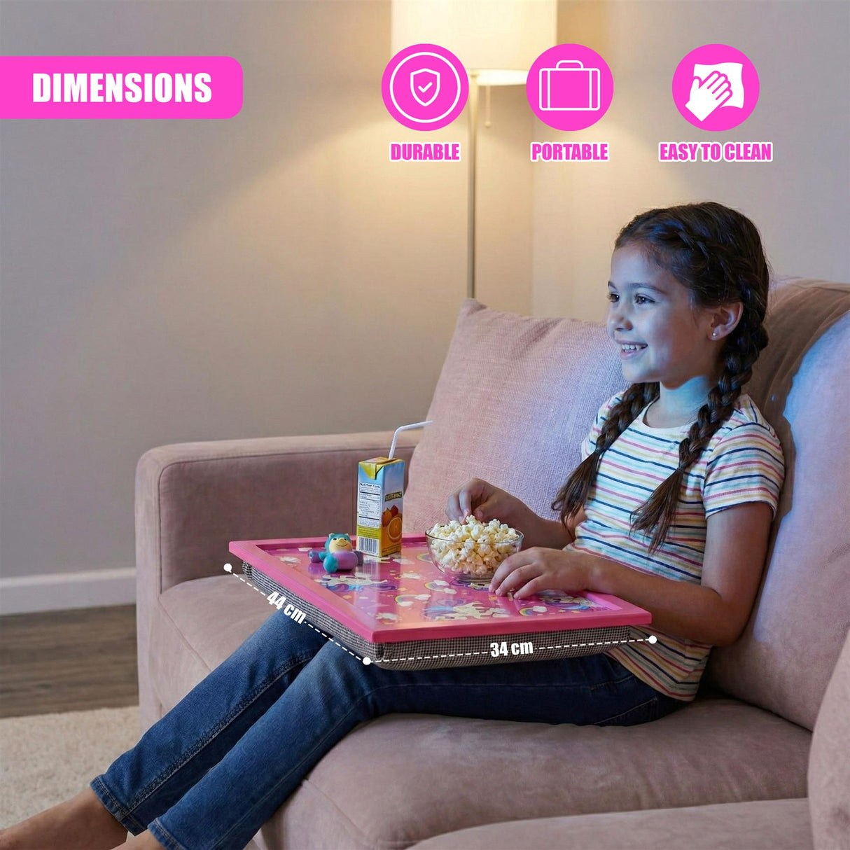 A young girl sits on a couch using a rectangular pink lap tray and eating snacks on her lap.