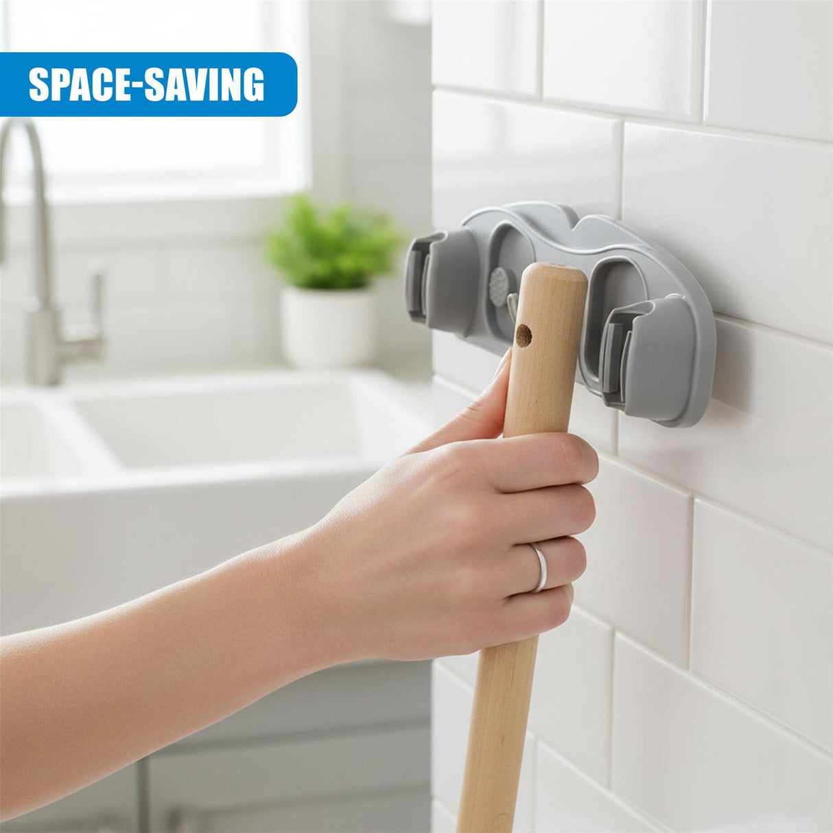 A hand holds a wooden mop handle next to a gray, wall-mounted cleaning tool holder.