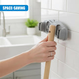 A hand holds a wooden mop handle next to a gray, wall-mounted cleaning tool holder.