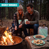 Couple enjoying snacks and eating off a green picnic set near a campfire in a wooded outdoor setting.