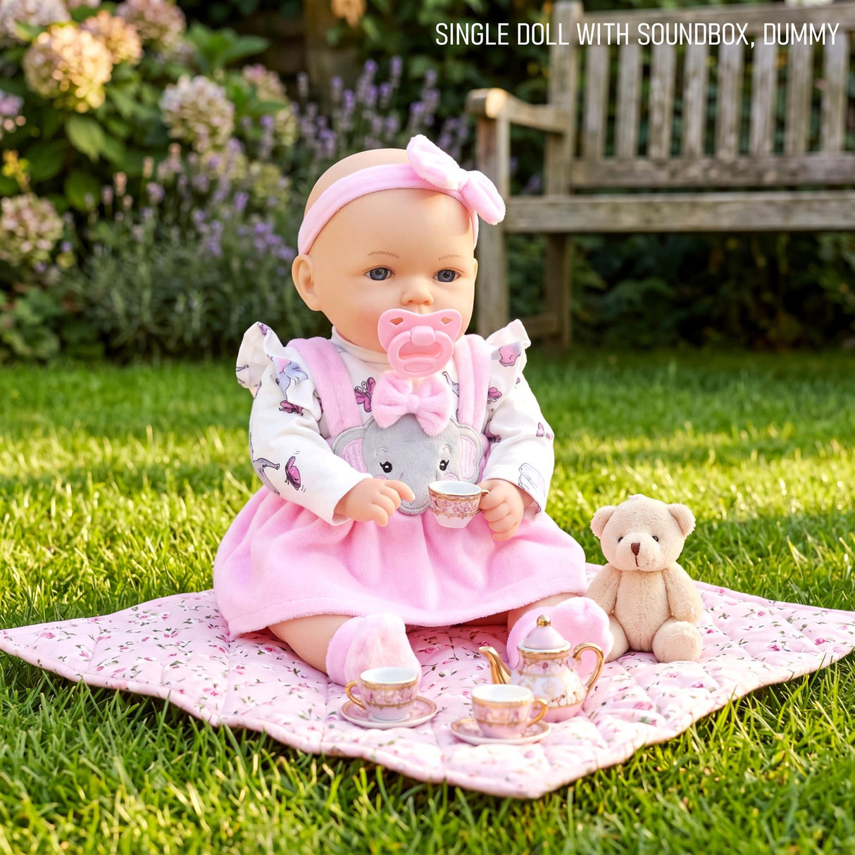 Pink doll, teddy bear, and teacup set arranged on a patterned blanket on green grass.