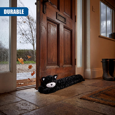 Fuzzy black cat draught excluder placed on the stone floor next to a wooden door.