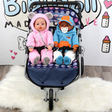 Two dolls dressed in pink and blue sit in a dark stroller on a fluffy white surface.