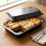 A dark baking pan holding cheesy casserole, with a fitted metal lid and forks nearby.