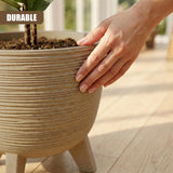 Large, ribbed brown flower pot on legs contains soil and a small plant. A hand rests near the rim.