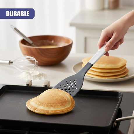 Spatula with holes rests on a griddle pan holding a round, golden pancake.