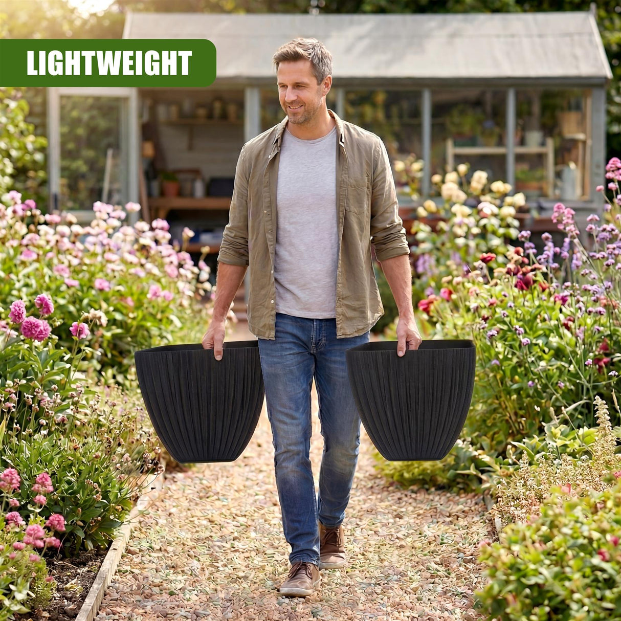 Man in a garden carries two black, textured, square planters next to blooming flowers.