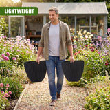 Man in a garden carries two black, textured, square planters next to blooming flowers.