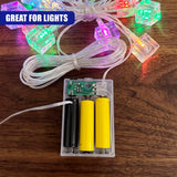 A clear plastic battery eliminator for AA and AAA batteries sits on a wooden surface, connected to a string of glowing LED