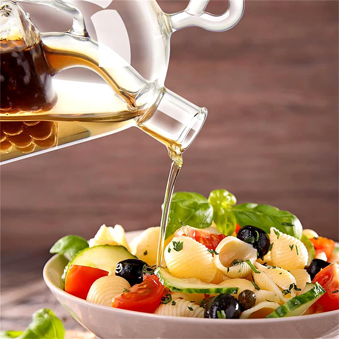 A glass oil and vinegar dispenser pouring liquid over a fresh pasta salad filled with tomatoes, olives, cucumber, and basil. The background features warm wooden tones, enhancing the appetizing appearance of the dish. Perfect for kitchen use and entertaining. Available at ukbuyzone.