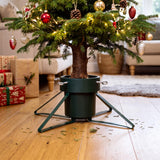 A green metal Christmas tree stand holding a decorated tree on a wooden floor with gifts nearby