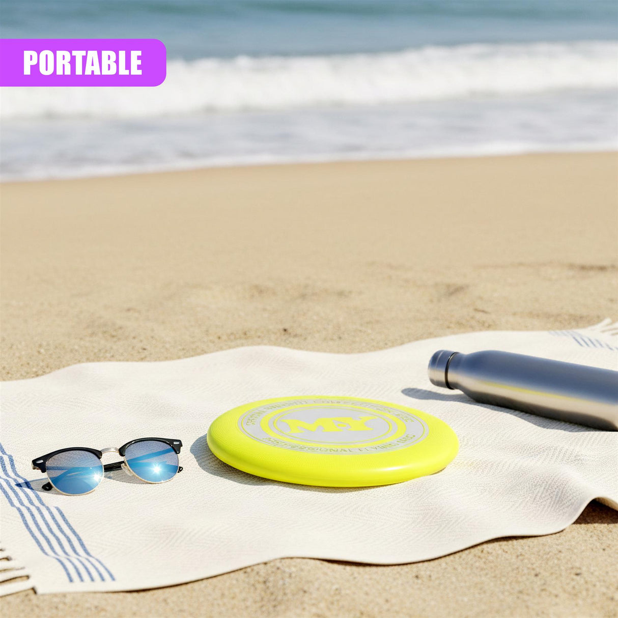 Yellow frisbee, sunglasses, and dark bottle rest on a white beach towel on the sand.